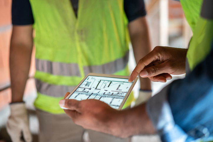Close-up of a male architect using a digital tablet for discussing construction plan with colleagues at a construction site. Construction site engineer reviewing blueprints on digital tablet.