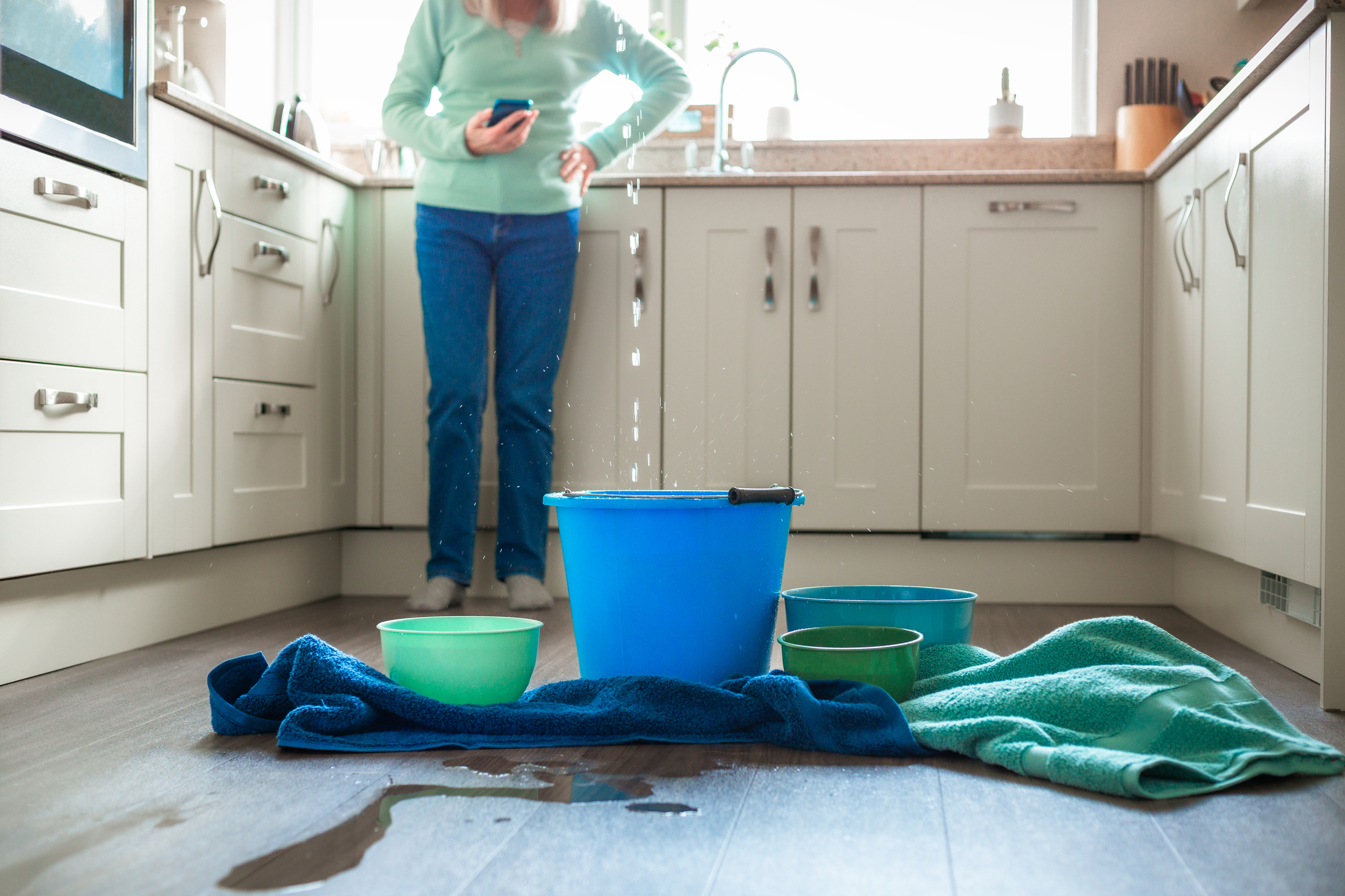 A senior woman looks worried as he deals with a water leak in the kitchen ceiling. Buckets and pots are on the floor to catch the flowing water, and towels are soaking up the puddles. The woman is defocused in the background, using his smart phone.